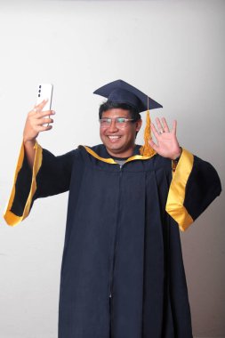 Mezuniyetini kutlamak için kep ve cübbe giymiş genç bir adam cep telefonunu selfie çekmek, arama yapmak ve baloda mesaj göndermek için kullanıyor.