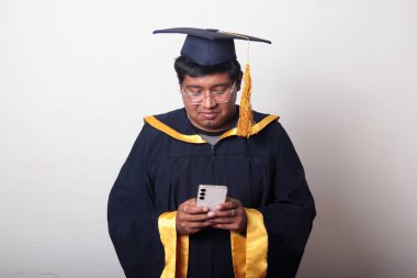 Mezuniyetini kutlamak için kep ve cübbe giymiş genç bir adam cep telefonunu selfie çekmek, arama yapmak ve baloda mesaj göndermek için kullanıyor.