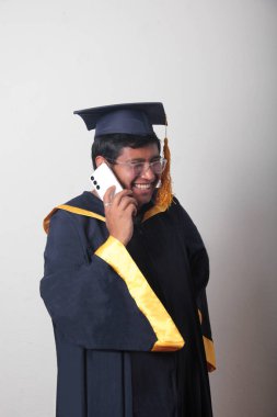 Mezuniyetini kutlamak için kep ve cübbe giymiş genç bir adam cep telefonunu selfie çekmek, arama yapmak ve baloda mesaj göndermek için kullanıyor.