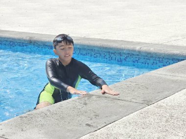 Latin çocuk tatilde ya da hafta sonu havuzda yalnız oynuyor. Kendini güneşten korumak için mayo giyiyor.