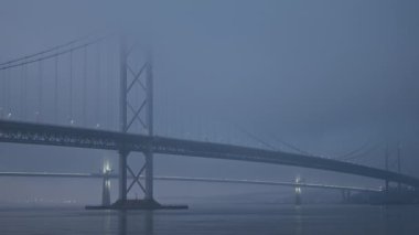 A large suspension bridge over the sea bay covered with fog or haar. The lights of cars moving across the bridge. Video with the panning camera motion. Forth Road Bridge, Scotland, United Kingdom