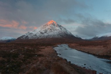 Landscape of the snowcapped Buachaille Etive Mor mountain and the Coe River at sunrise.The rising sun only illuminates the top of the mountain. Scottish Highlands, UK. 