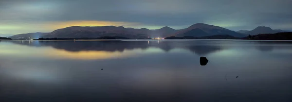 Panorama of a lake and mountains at night. The sky illuminated by the street lights of the towns. Loch Lomond, Scotland