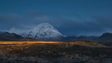 Glen Coe 'nun İskoç Highlands' ına giden bir yolun gece görüşü. Araba ışık yolu, karlı dağlar ve vadi. İskoçya