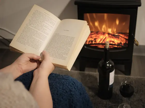 Woman reading book in front of the fireplace. There is a glass of wine and a bottle on the floor. Only the womans hands are visible. Winter and long cold night concept. Close up, selective focus