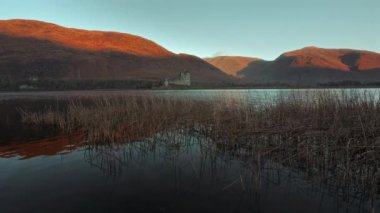Sazlıklarla dolu bir gölün kıyısında duran antik bir şatonun İskoç kalıntıları. Dağlar, sabah güneşinin ışınlarıyla aydınlanır. Kilchurn Kalesi, Loch Awe, Argyll ve Bute, İskoçya, İngiltere