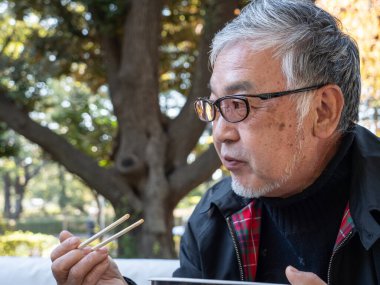 Yaşlı bir Japon adam dışarıda çubuklarla sıcak erişte ve çorba yiyor.
