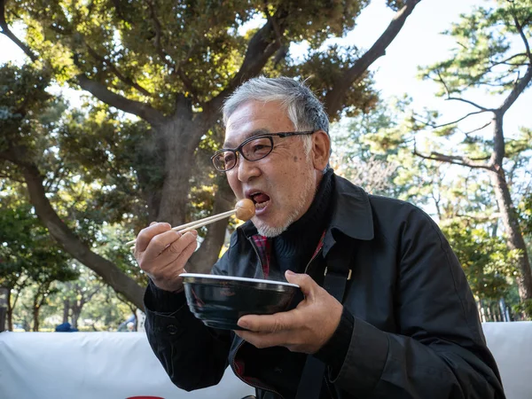 An older Japanese man eating warm noodles and soup with chopsticks outside