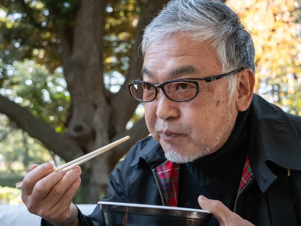 An older Japanese man eating warm noodles and soup with chopsticks outside