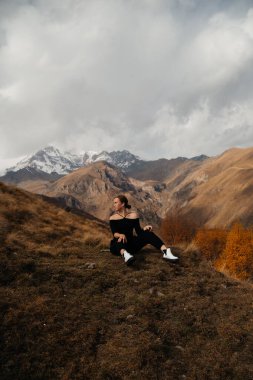 Tepede oturan genç ve güzel bir kadın. Arka planda kar örtülü dağlar. 