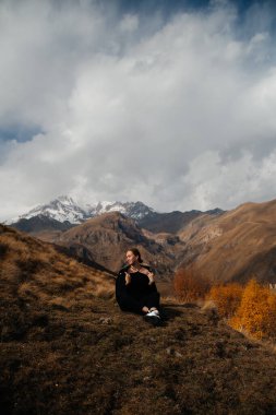 Tepede oturan genç ve güzel bir kadın. Arka planda kar örtülü dağlar. 