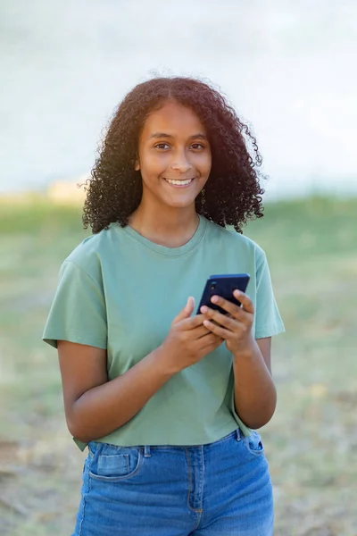 夏の日の間にスマートフォンを使用して巻き毛を持つ美しい笑顔の女の子 — ストック写真