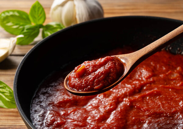 Scoop up the tomato sauce in the pan with a spoon. Cooking scene. Close-up. Wooden background. Basil and garlic in background.