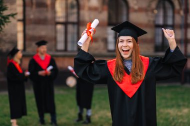 Uzun, düz saçlı, mutlu genç bir kadın, ellerini kaldırıyor, kırmızı kurdeleli bir diploma tutuyor üniversiteden mezun olduğu için mutlu..