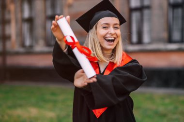 Üniversitenin arka planında tek başına duran bir öğrenci, mutlu bir şekilde diplomasını gösterir ve eğitiminin sonunu kutlar..