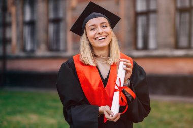 Üniversite bahçesinde elinde diplomayla bekleyen mutlu bir mezun kameraya bakıp gülümsüyor..