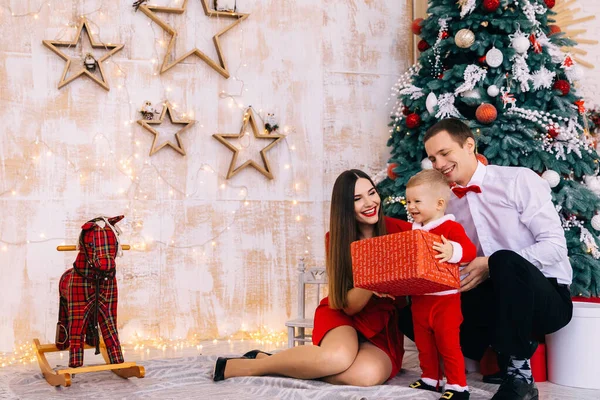 サンタの衣装の赤ちゃんは大きなギフトボックスと笑顔を保持します 両親はクリスマスツリーの近くに座って笑っていました 部屋に馬を揺らし — ストック写真
