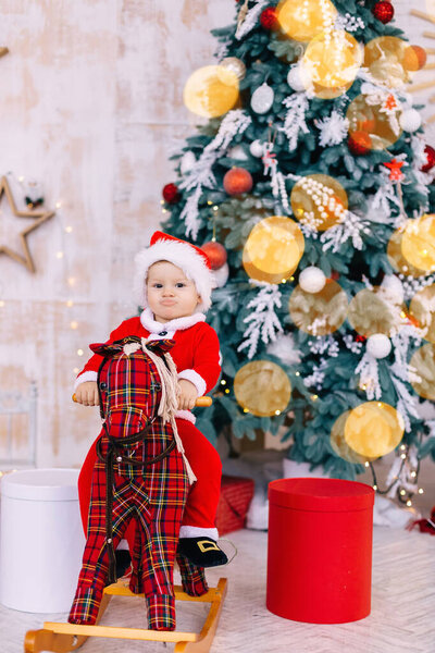 little boy in santa costume on rocking horse look at camera on the background of Christmas tree and gifts.