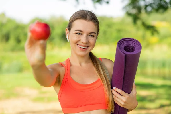 スポーツウェアを着た美しい若い女性がヨガマットと赤いリンゴを持っていて カメラを見て笑っていて 公園に立っています — ストック写真