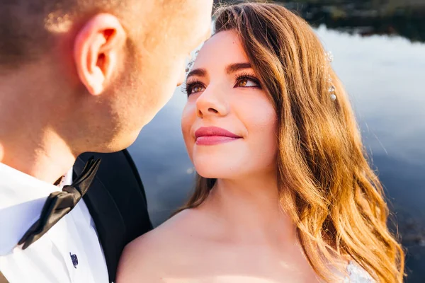 Close-up of a bride with a beautiful makeup and a hairdo. the bride looks at the groom. groom in a suit with bow tie. evening lake
