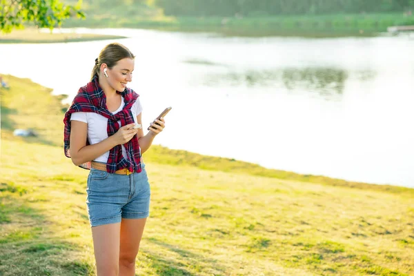 Mutlu bir kızın portresi, güneşli bir yaz gününde, sokakta bulunan akıllı bir telefondan kablosuz kulaklıkla müzik dinliyor.
