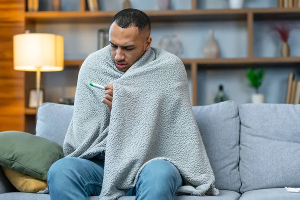 Man wrapped in warm knitted plaid trembling with cold sit alone on sofa. Central heat problems, influenza symptoms concept.