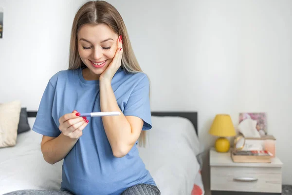 若い女性が妊娠検査を笑顔を保持ベッドの上に座って肯定的な2本の縞模様の結果を参照してください 健康な女性排卵期間妊娠の概念を検出するための現代的な装置 — ストック写真