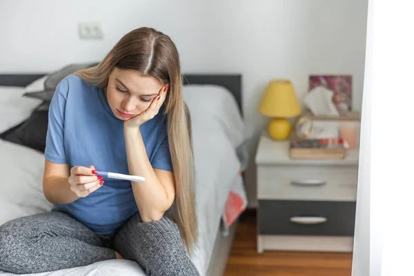 Upset blonde girl checking her recent pregnancy test, sitting on bed at home, copy space
