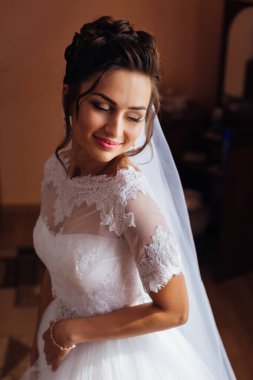 Hoş bir gelin, uzun bir gelinlik giymiş, dantelli ve saçında peçesi olan bir kadın törene hazırlanıyor.