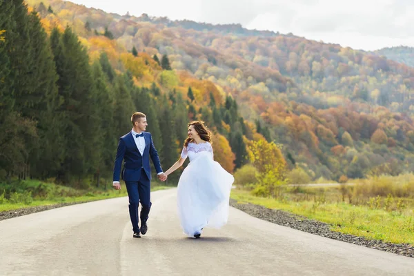 一位身穿白衣的快乐的新娘与新郎手牵着手 在美丽的大自然的衬托下沿着大路跑着 — 图库照片