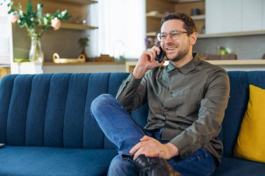 Yakışıklı genç adam akıllı telefonda konuşuyor ve kanepede otururken gülümsüyor.