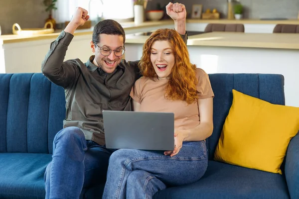 Shocked young family couple cuddle on sofa raise hands scream wow read email with great news on laptop screen. Lucky husband wife celebrate winning lottery getting great offer proposal for purchase