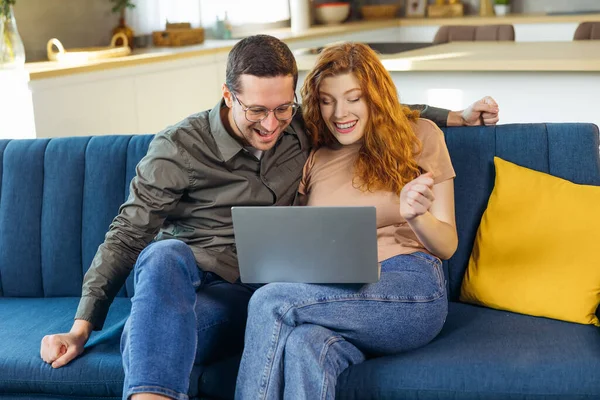 Overjoyed couple sit relax on cozy couch look at laptop screen triumph winning lottery online, happy biracial husband and wife feel excited euphoric with good news on computer
