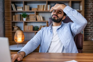 Yorgun Arap ya da Hintli adam, ofis çalışanı, yönetici ya da serbest çalışan, masasında oturan, laptopta çalışmaktan yorgun, çok çalışan, baş ağrısı çeken, gözlerini kapayan, dinlenmeye ihtiyacı olan