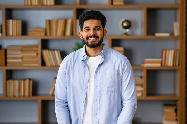 Smiling bearded indian businessman looks at the camera. Young positive male student in library with bookshelves on background. Proud and successful mixed-race small business owner