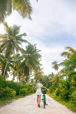 Hasır şapkalı kadın eski model bisikletle palmiye ağaçları arasında yürüyor.