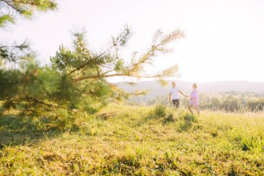 Genç ve güzel giyimli hamile bir kadın akşam yürüyüşü sırasında bir tarlada kocasının elini tutuyor. Hamilelik ve bakım. Mutluluk ve şefkat. dikkat ve dikkat