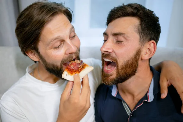 stock image guys sitting on the couch in a modern apartment. Gay couple are having lunch, eating pizza. The couple talks and enjoys being together. LGBT couple concept at home