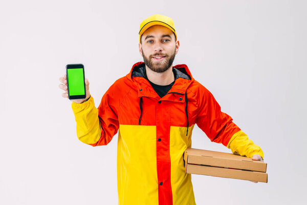 A courier in a red and yellow jacket and a cap, hold pizza boxes in one hand, and a phone with a green screen in the other. Advertising of the application for free delivery with low prices for fast food.