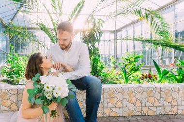Gelin ve damat bir botanik bahçesinde, yeşilliğin arka planında oturuyorlar.