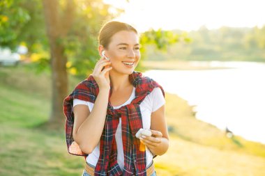 Telefonu elinde tutan ve gülümseyen bir kız. Kablosuz kulaklıklardan müzik dinlemek. Güneş parlıyor. Gün batımı.
