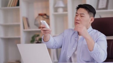 Dizüstü bilgisayarda çalışan Asyalı işadamı, klima soğutma sistemindeki uzaktan kumandayı ya da ofisteki bilgisayar masasındaki nemlendiriciyi çalıştırıyor.