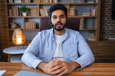 Kamerayla konuşan Hintli erkek öğretmen çevrimiçi ders veriyor oturma odasında oturuyor evdeki kitaplığın yanında. Uzaktan Öğretmenlik ve E-Öğretim Konsepti. Seçici Odaklanma