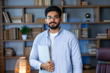 Smiling IT worker in blue shirt look at camera holding laptop at office. concept of remote and freelance work. millennial successful man in glasses clipart