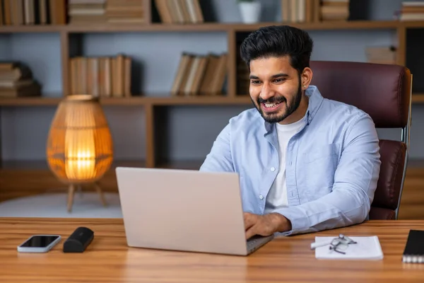 Portrait Of Smiling Middle Eastern Male Manager Working On Laptop Computer In Modern Office, Cheerful Guy Sitting At Desk And Using Pc, Looking At Screen Typing On Keyboard, Enjoying Remote Job
