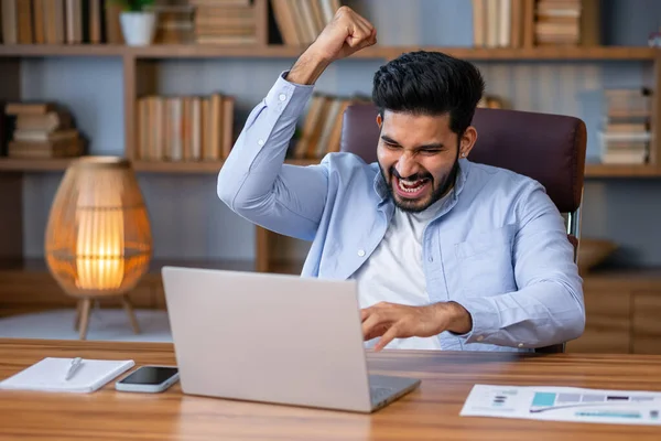 Overjoyed happy indian guy, lucky winner, sitting at desk watching virtual lottery results on laptop shaking hands. Eastern student got perfect exam grades, job proposal offer.