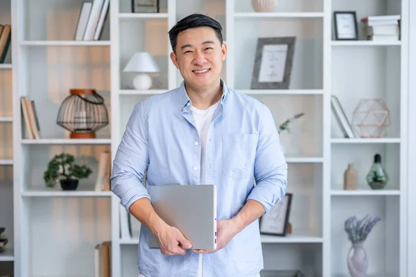 stock image Confident young asian man in casual shirt holding laptop while standing in home office