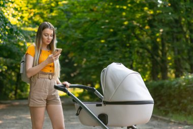 Anne, bebek arabasında elinde telefonla bebeği sallıyor. Kadın, yeşil ağaçların yakınındaki parkta postaları kontrol ediyor.