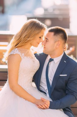 Yeni evliler bir bankta oturuyorlar gözleri kapalı ve gülümsüyorlar. Şehirde güneşli bir gün.