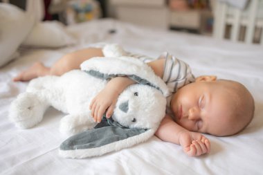 Huzurlu bir bebek, çarşafların üzerinde yatıyor oyuncakla sarılıp uyuyor